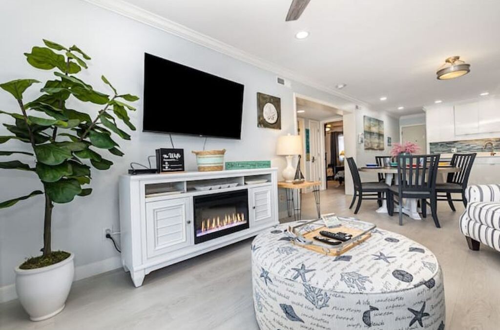 A modern living room with a flat-screen TV, a fireplace, a dining area with a table and chairs, and a potted plant.