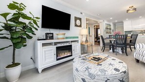 A modern living room with a flat-screen TV, a fireplace, a dining area with a table and chairs, and a potted plant. - The Sea Cabin w/ Partial Ocean View & Pool, Golf, Beach (North Myrtle Beach)