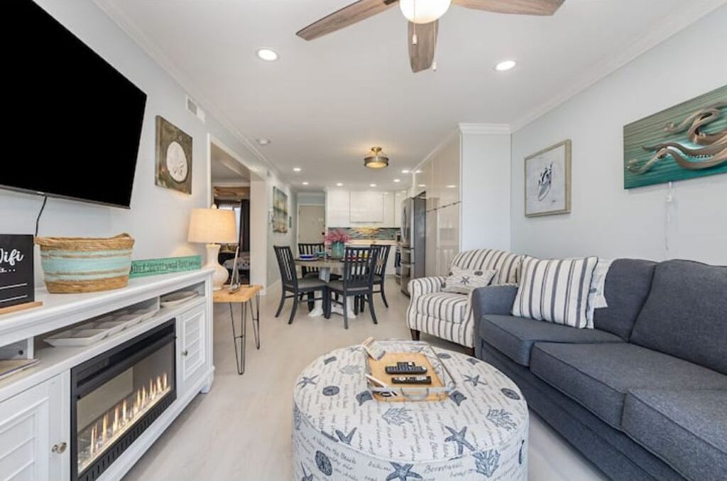 A living room with a grey sofa, a striped armchair, a round ottoman with nautical designs, a flat-screen TV, a fireplace, and a dining area with a table and chairs.