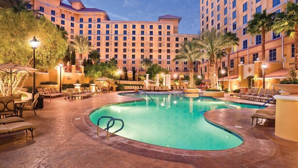 A hotel pool area with lounge chairs and umbrellas, surrounded by palm trees and a large building in the background.