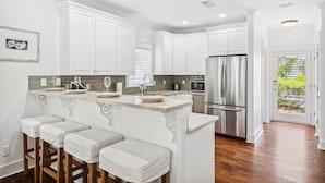 A modern kitchen with white cabinetry, a central island with bar stools, stainless steel appliances, and a view to the outdoors. - 30A Beach House - Beach Bikin (Panama City Beach)