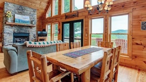 Interior de una cabaña de madera con chimenea de piedra, un sofá, una mesa de comedor con sillas y ventanas amplias con vistas al exterior.