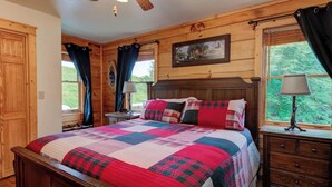 Un dormitorio de cabaña de madera con una cama grande, dos mesitas de noche, un ventilador de techo y una ventana con cortinas.