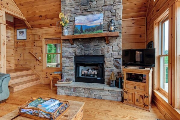 A cozy cabin interior with a stone fireplace, wooden walls, a television, and a bookshelf. - Wildlife Ridge-Views, Gameroom, Hot Tub 4300' Elev (Mars Hill)