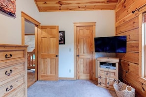 A room with wooden walls and ceiling, a flat-screen TV mounted on the wall, a wooden cabinet with drawers, and a door leading to another room.