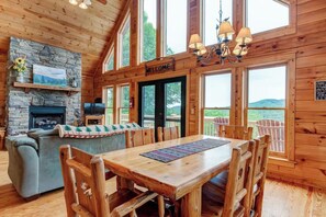 A wooden cabin interior with a stone fireplace, a sofa, a dining table with chairs, and large windows offering a view of the outdoors.