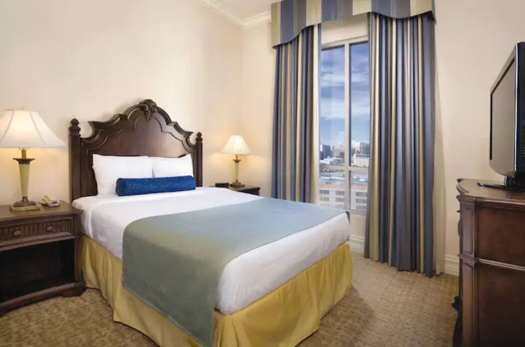 A hotel room with a large bed, two bedside tables with lamps, a television on a dresser, and a view of the city through the window.
