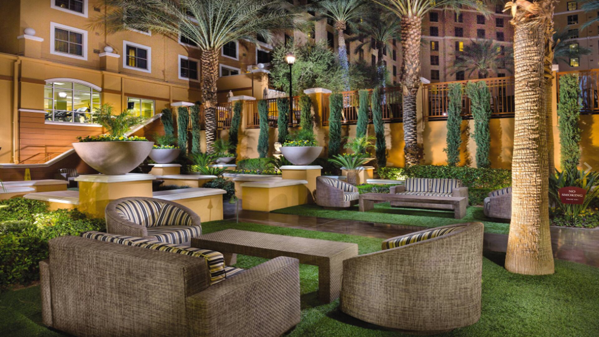 An outdoor lounge area with sofas and planters, surrounded by palm trees and greenery.