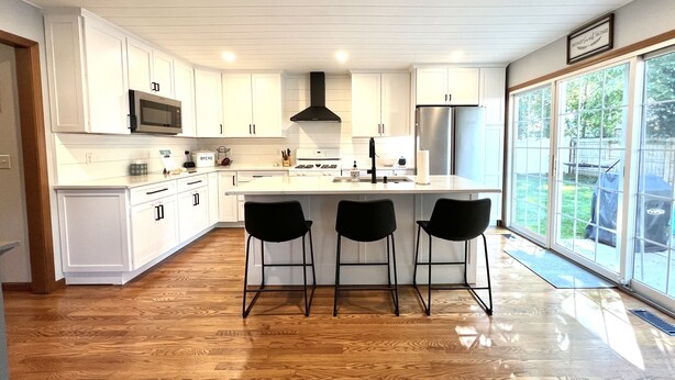 A modern kitchen with white cabinetry, a central island with bar stools, a stainless steel refrigerator, and a large window offering a view of the outdoors. - ☘️ 10 mins to Notre Dame, 4 Bedrooms, Garage Park, Restaurants Close. Go Irish!☘️ (South Bend)