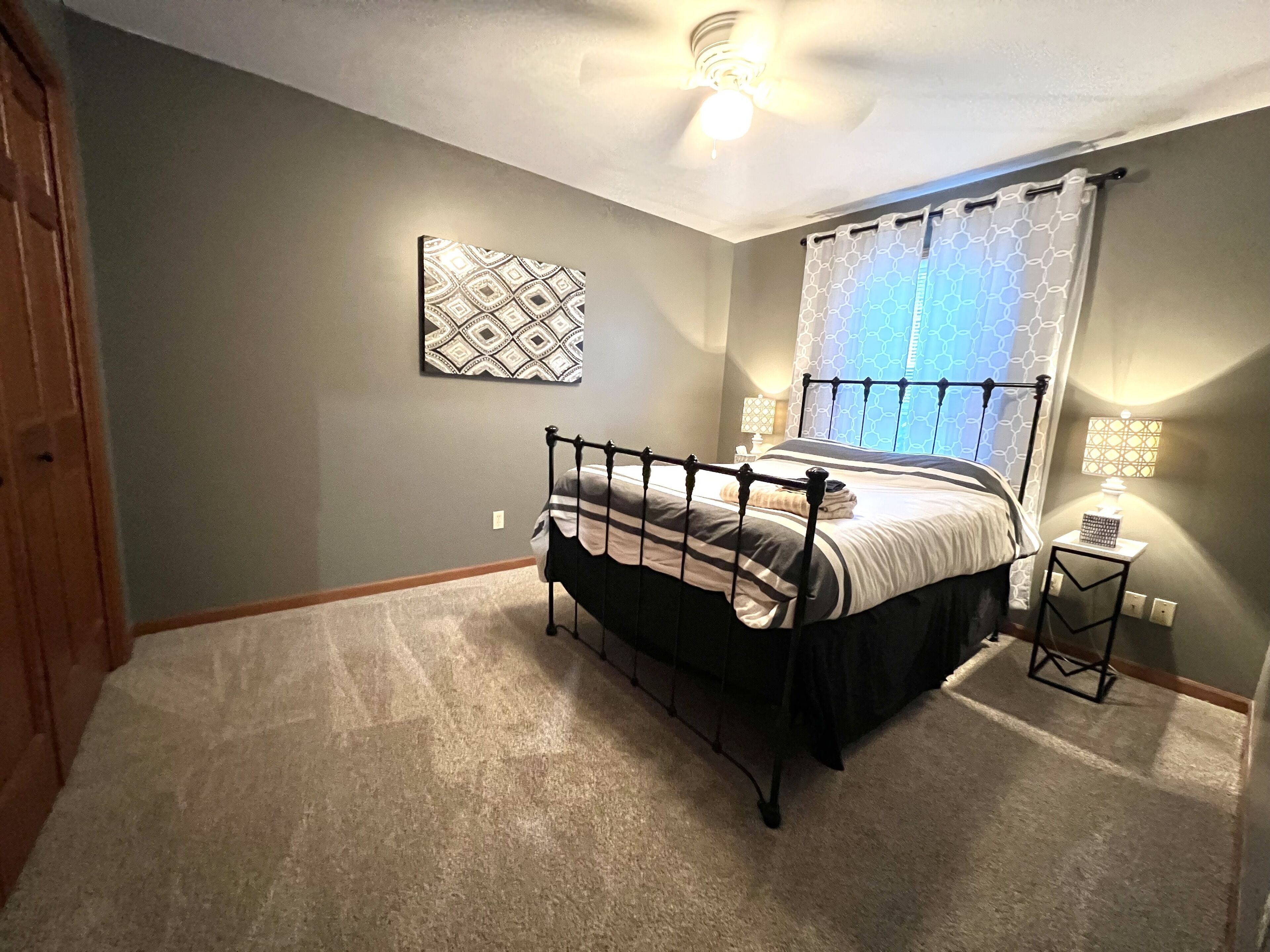 A bedroom with a bed, a ceiling fan, a wall-mounted lamp, and a window with a curtain.