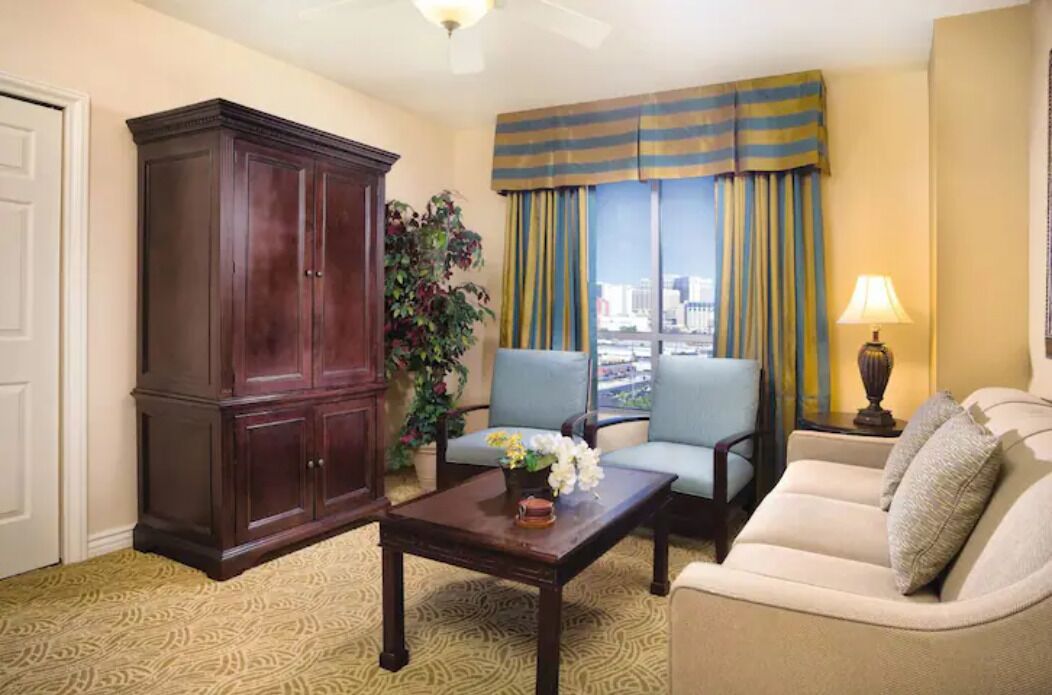 Una habitación con un armario grande de madera oscura, un sillón azul, un sofá beige y una mesita con una lámpara.