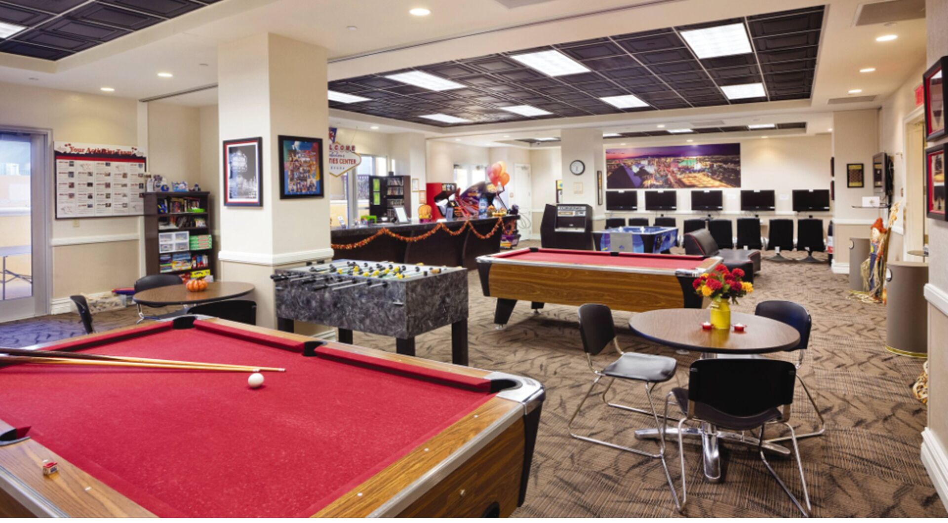 A pool table, foosball table, and a billiards table in a room with multiple screens and a clock.