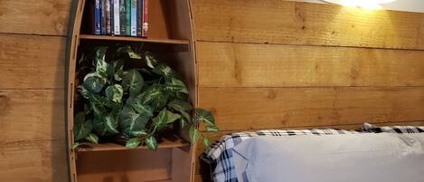 Quarto com cabeceira de madeira, estante com livros e uma planta, cama com roupa de cama branca e luminária fixada na parede.