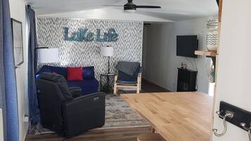 Una sala de estar moderna con sofá, una mesa de centro y un televisor. Hay una mesa de comedor de madera con taburetes.