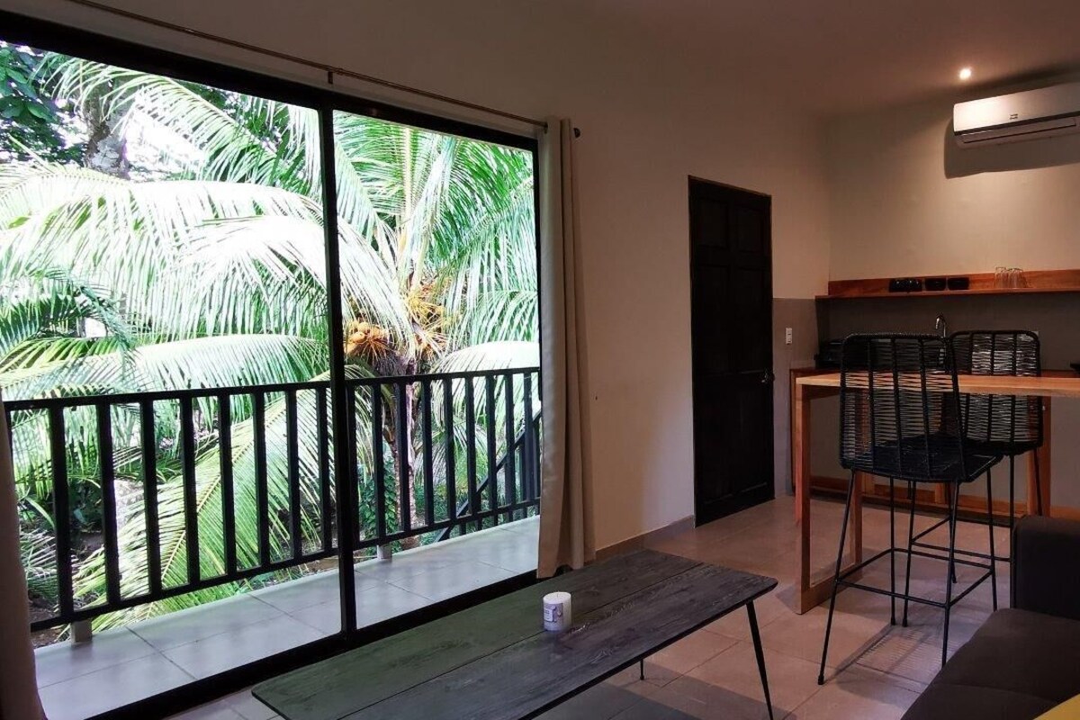 Varanda com vista para palmeiras, uma mesa pequena com uma xícara, um sofá e uma cozinha com bancada e cadeiras.