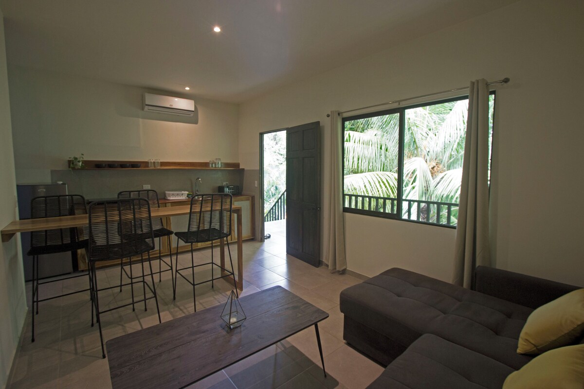 Uma sala de estar moderna com sofá, área de jantar com cadeiras e uma mesa, e uma janela grande com vista para o verde.