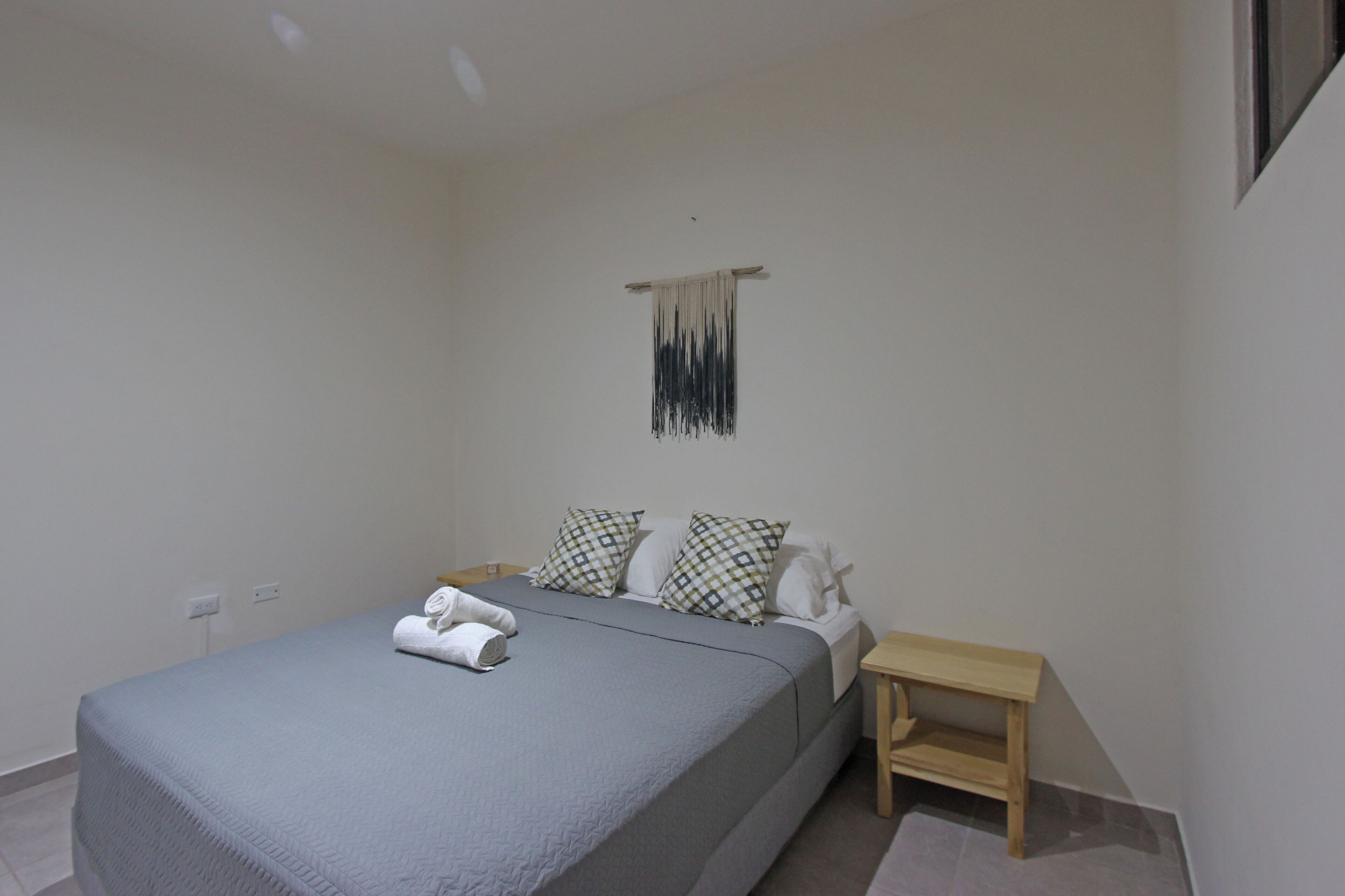 A neatly made bed with a grey bedspread and white pillows, a small wooden nightstand, and a wall-mounted hanging.