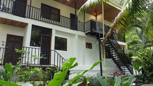 A two-story building with a balcony, a staircase, and lush tropical vegetation.