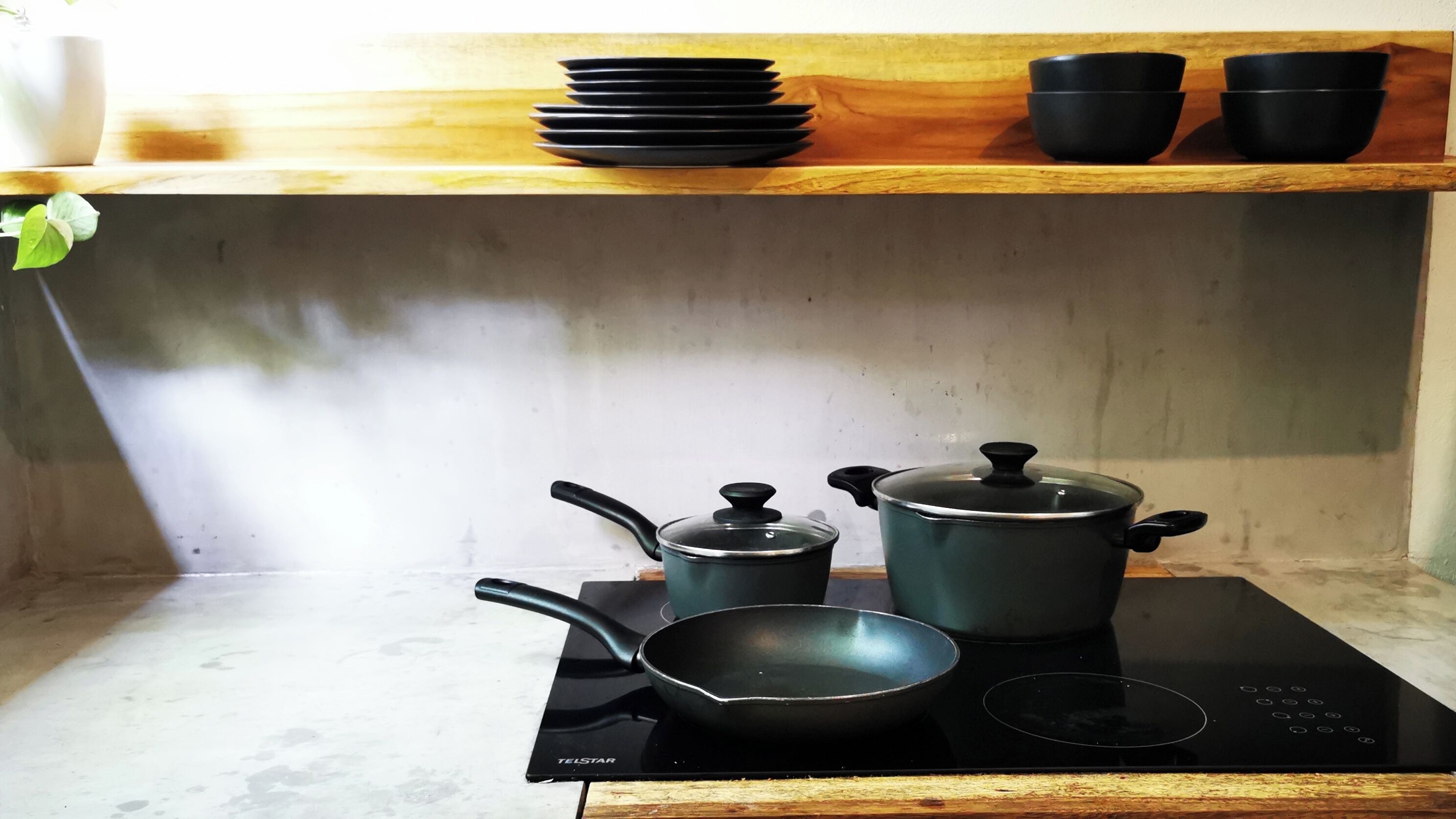 Una cocina con estufa, sartén, olla y una pila de cuencos negros sobre un estante de madera.