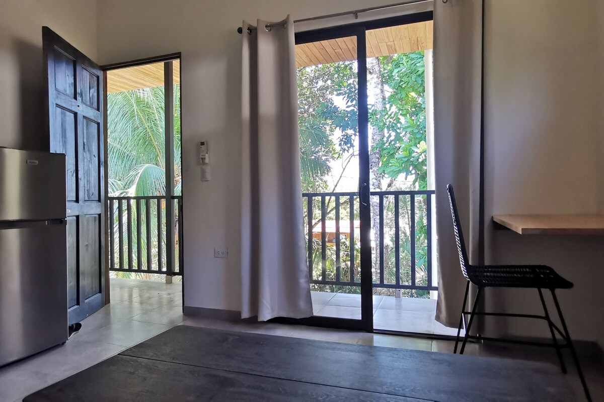 Quarto com porta de vidro deslizante para uma varanda, geladeira e uma pequena mesa com uma cadeira.