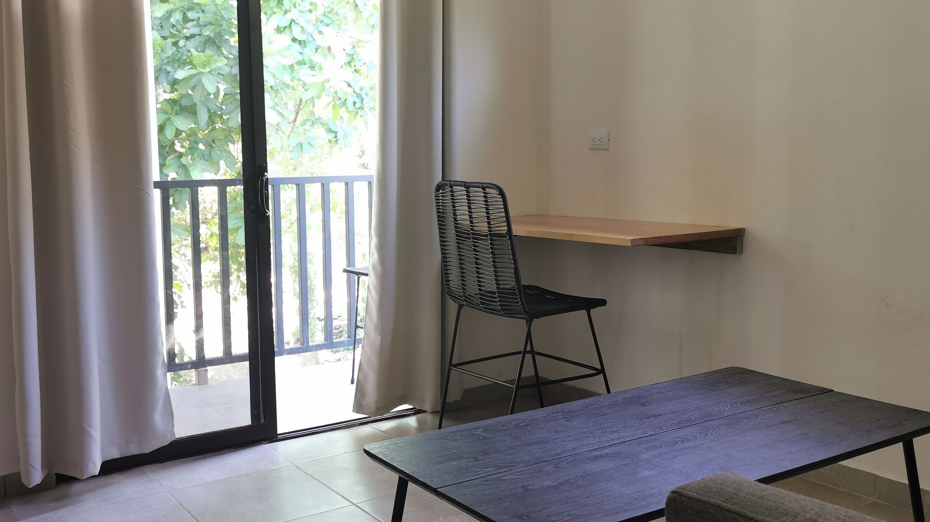 A room with a sliding glass door leading to a balcony, a black metal chair, a wooden table, and a wall-mounted shelf.