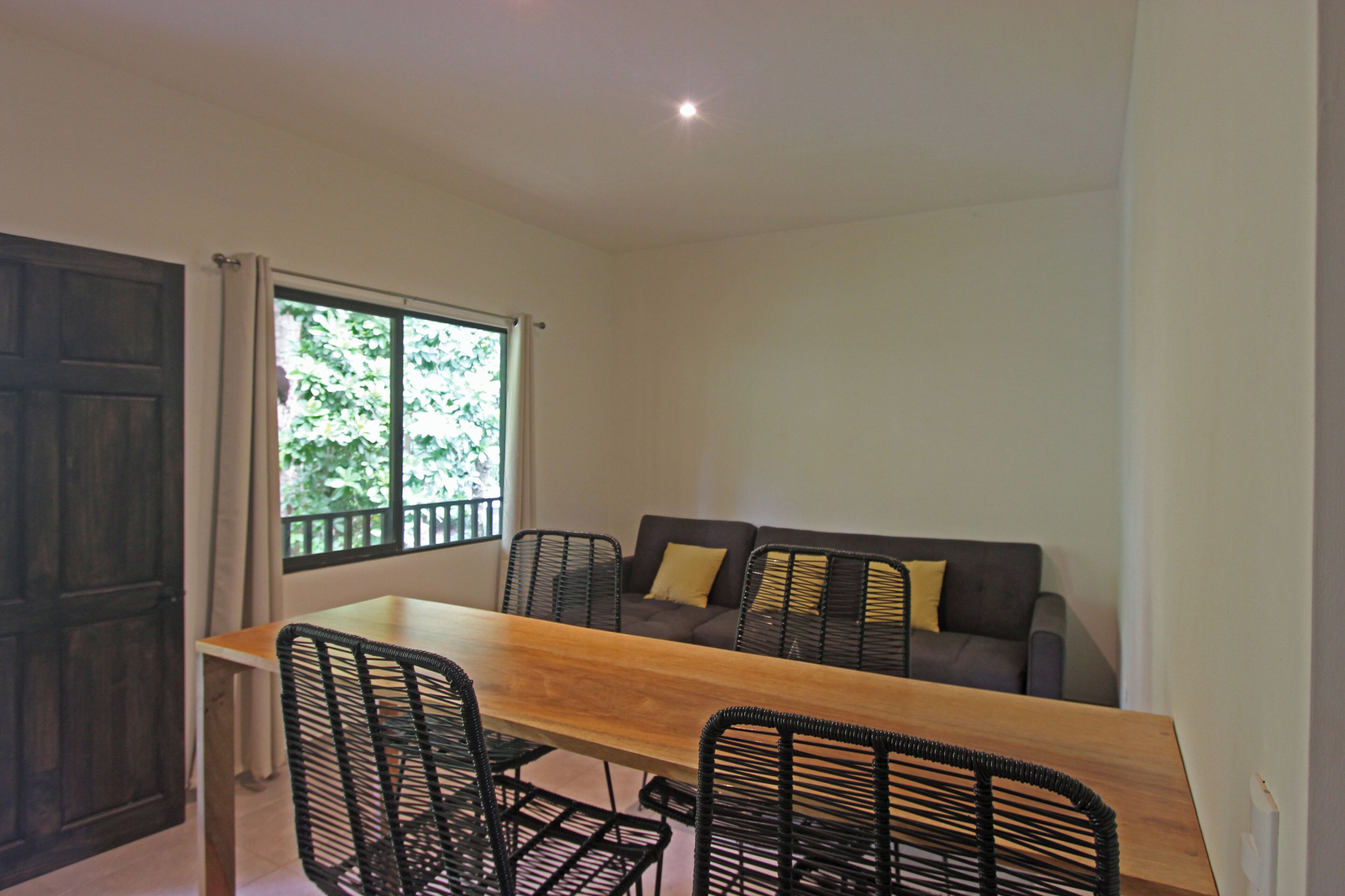 A dining area with a wooden table and black chairs, a sofa with cushions, and a window with greenery outside.
