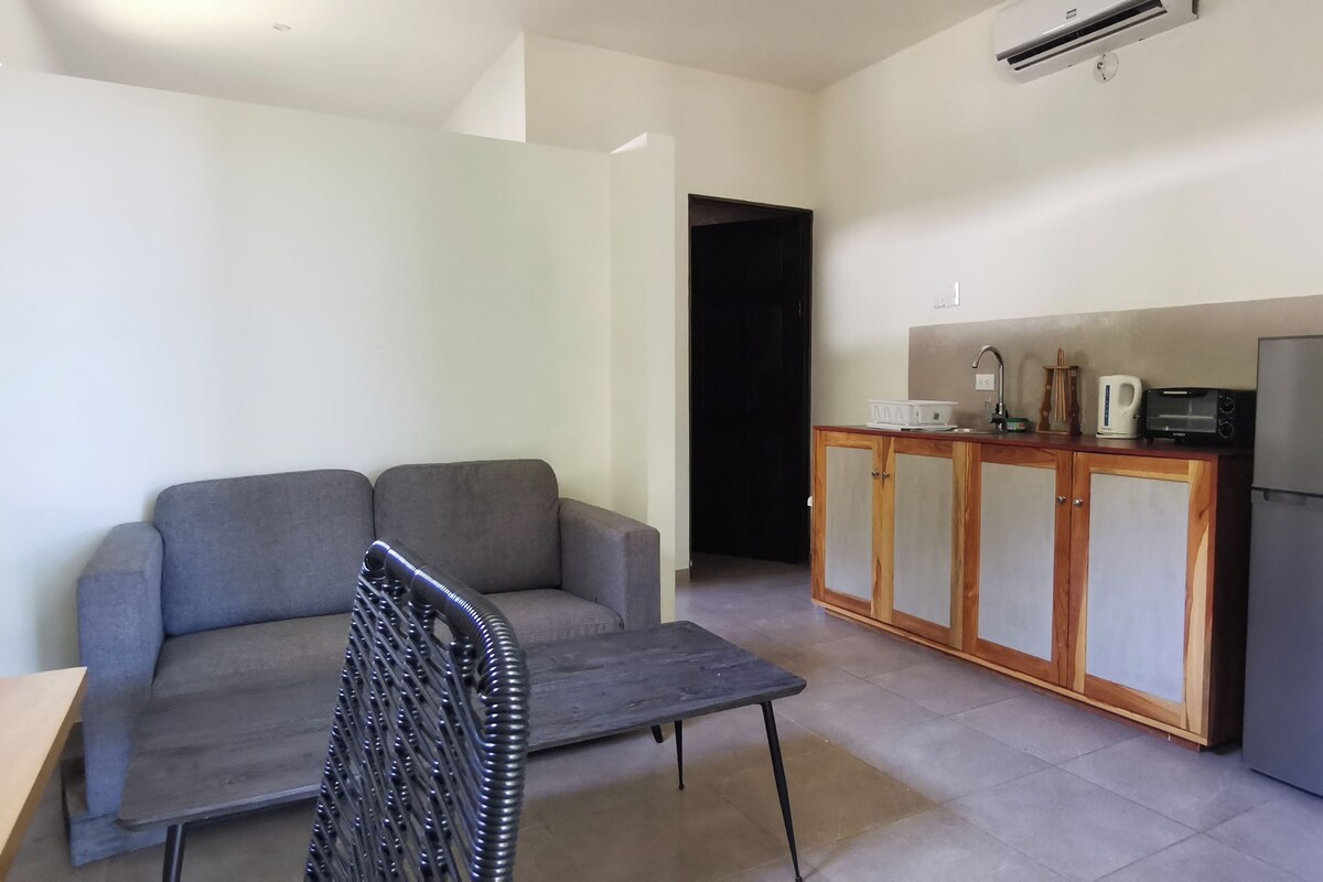Um espaço de estar compacto com sofá cinzento, uma mesa pequena, e uma cozinha compacta equipada com frigorífico, micro-ondas e pia.