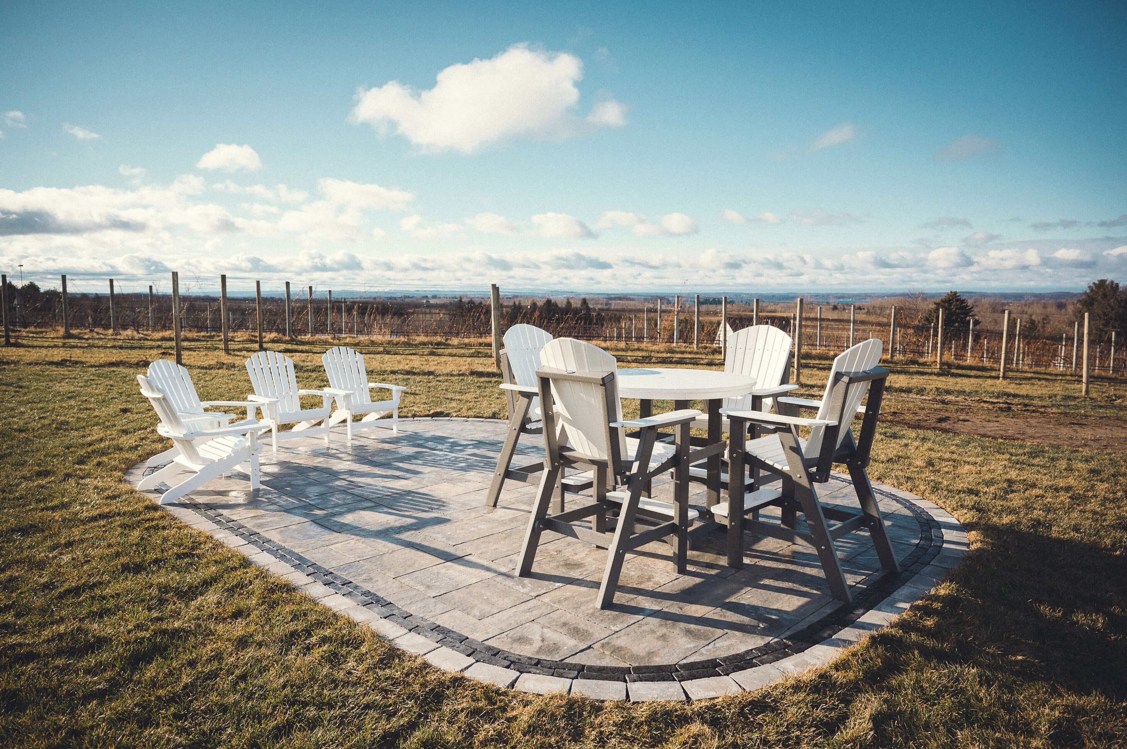 A patio with a table and chairs, surrounded by a brick border, set in a grassy field with a fence in the background.
