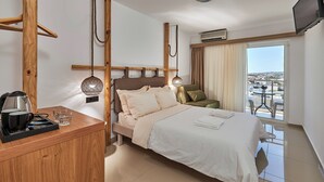 Habitación de hotel con una cama, un escritorio de madera con tetera y vasos, un televisor y un balcón con vistas.