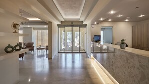 Un vestíbulo de hotel espacioso con un mostrador de recepción de mármol, un área de descanso con una mesa redonda y un amplio ventanal que da al exterior.
