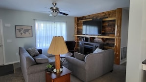 A living room with two grey sofas, a wooden coffee table, a ceiling fan, a flat-screen TV mounted on a wooden wall, and a world map on the wall. - Hillside Relaxation in Endless Mountains. Minutes from Tioga Downs. Sleeps 8 (Rome)