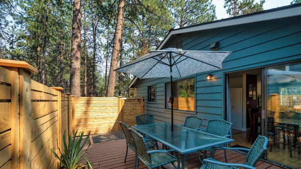 A deck with a glass table and chairs, surrounded by wooden fences and a house with large windows. - Westside Bend: Walk to Eateries & Downtown! (Bend)