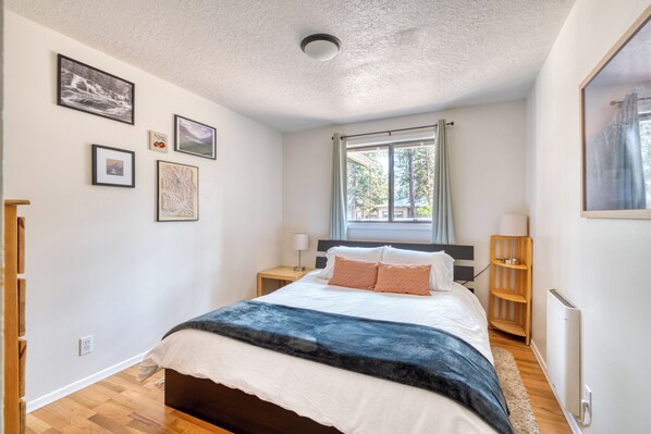 A bedroom with a bed, a window with curtains, a bedside table, and a wall with framed pictures.
