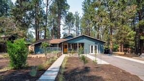 A single-story house with a front porch, surrounded by a garden and tall pine trees. - Westside Bend: Walk to Eateries & Downtown! (Bend)