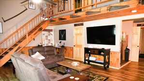 Ein geräumiges Wohnzimmer mit einer Holztreppe, einem großen Flachbildfernseher, einem Ledersofa und einem Couchtisch.