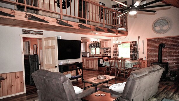 Ein gemütliches Wohnzimmer mit einer Couch, einem Couchtisch und einem Fernseher. Im Hintergrund ist eine Küche mit einem Esstisch und Stühlen zu sehen.
