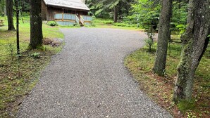 Ein Kiesweg führt zu einer rustikalen Holzhütte mit einem Metalldach und einer Holzterrasse, umgeben von dichtem Wald.