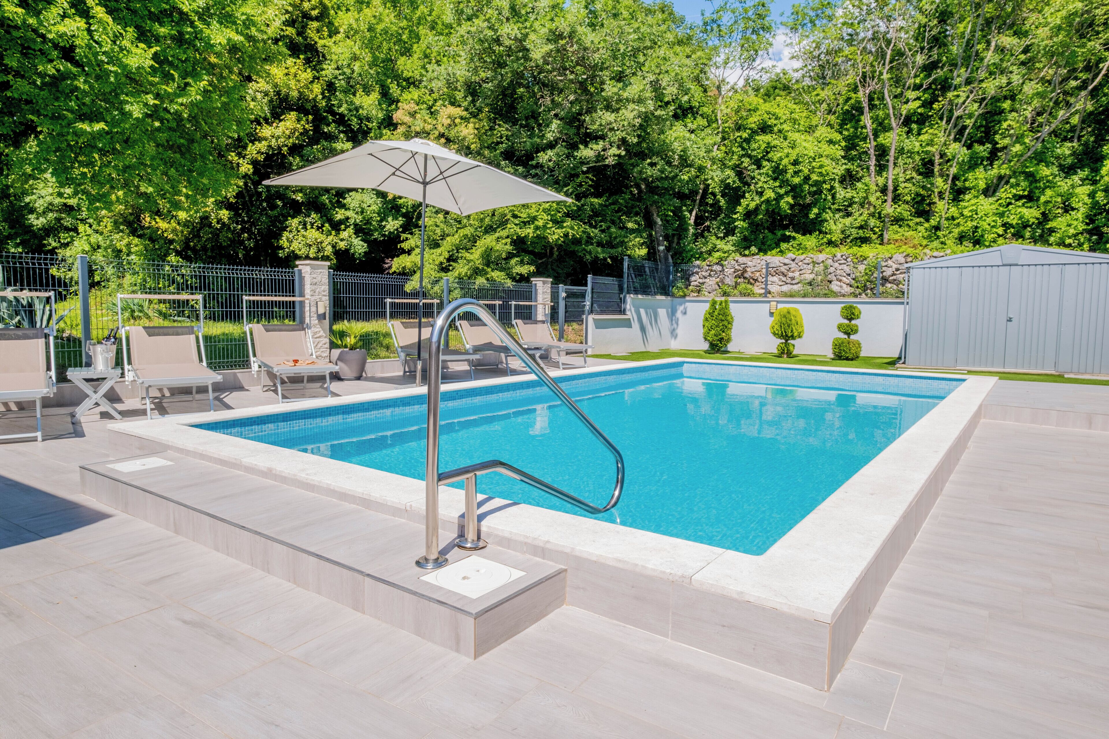 A rectangular swimming pool with a metal ladder and a white umbrella, surrounded by a fence and trees.