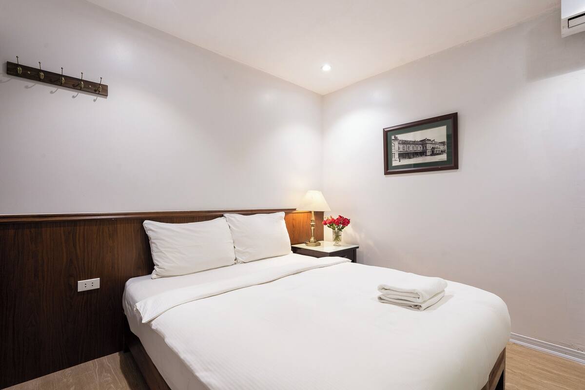 A neatly made bed with white linens, a bedside table with a lamp and flowers, and a framed picture on the wall.