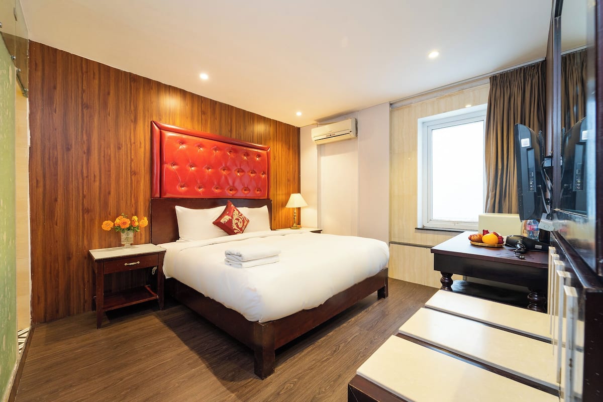 A hotel room with a wooden headboard, a bed with white linens, a bedside table with a lamp, and a TV on a stand.