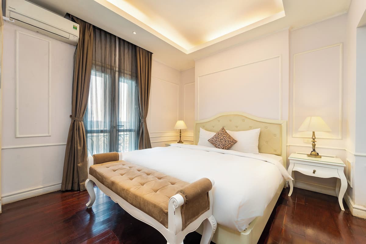 A hotel room with a large bed, a bench, two bedside tables with lamps, and an air conditioning unit.