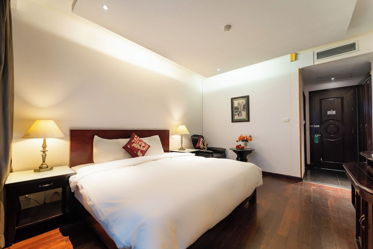A hotel room with a large bed, two bedside tables with lamps, a small table with a vase of flowers, and a door leading to another room.