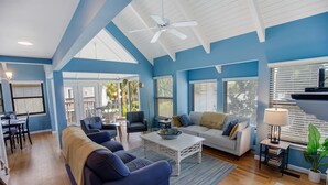 Uma sala de estar espaçosa com esquema de cores azul e branco, um ventilador de teto e uma área de jantar com mesa e cadeiras.