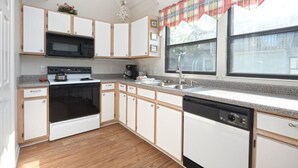 Una cocina con armarios blancos, microondas, estufa, lavavajillas y una ventana con cortina escocesa.