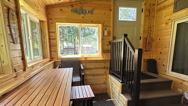 A wooden cabin interior with a wooden table and benches, a staircase, and a window with a view of trees.