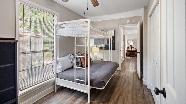 Une chambre avec un lit superposé, un ventilateur de plafond, une fenêtre avec des stores et un couloir menant à une autre pièce.