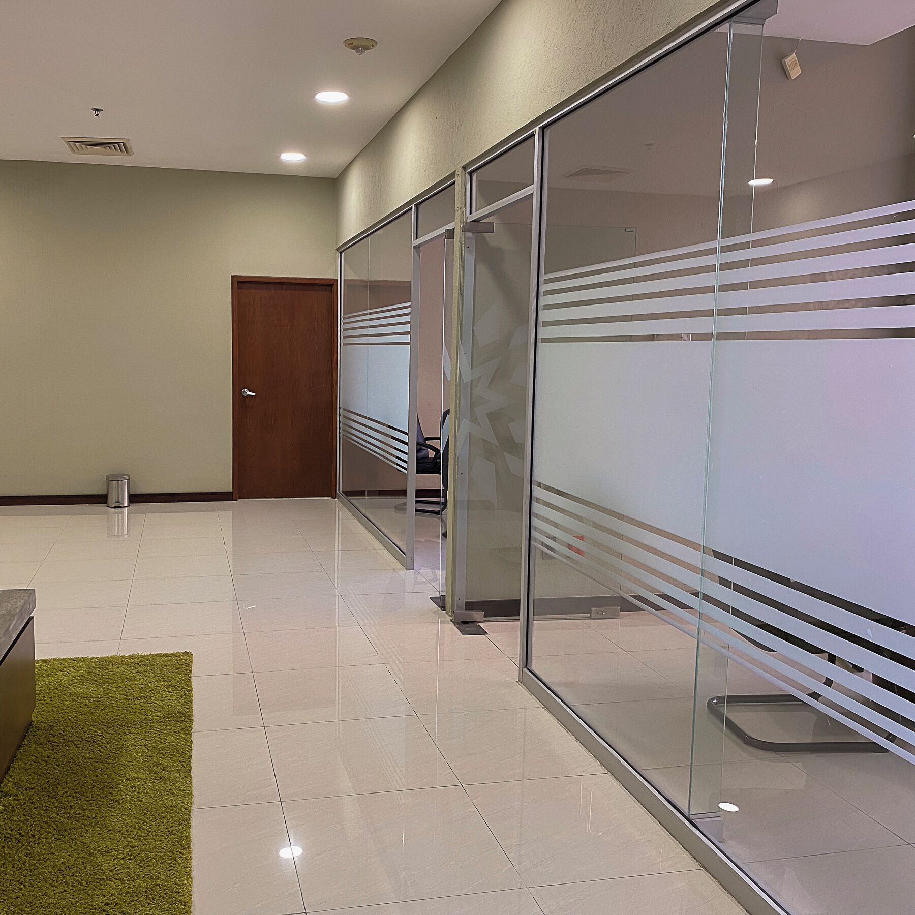 A hallway with glass partitions, a wooden door, and a trash can.