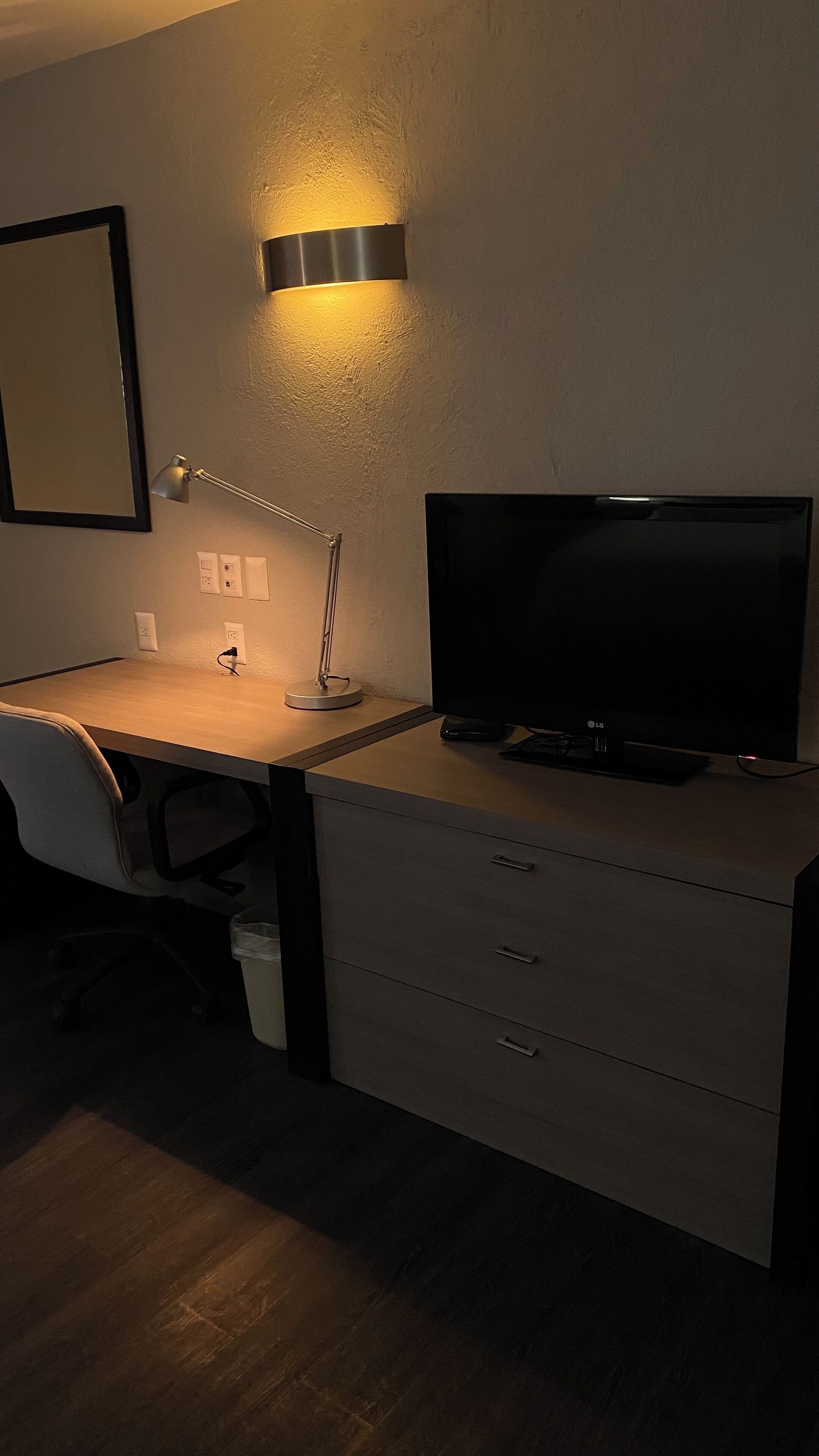 A hotel room with a desk, chair, TV, and a lamp.