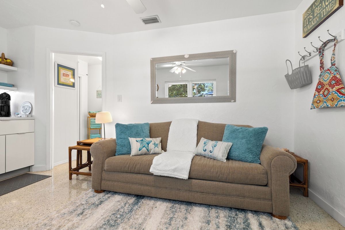 A living room with a beige sofa, a coffee table, and a rug. There is a kitchen area with a refrigerator and a coffee machine. A mirror hangs on the wall, and a basket hangs on a hook.