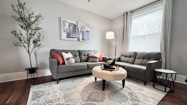 A living room with a grey sectional sofa, a round ottoman, a floor lamp, and a large window with curtains. - Spacious 3 Bedroom Sleeps 5 - Walk to Botanical Garden - Long Term Pet Friendly (St. Louis)